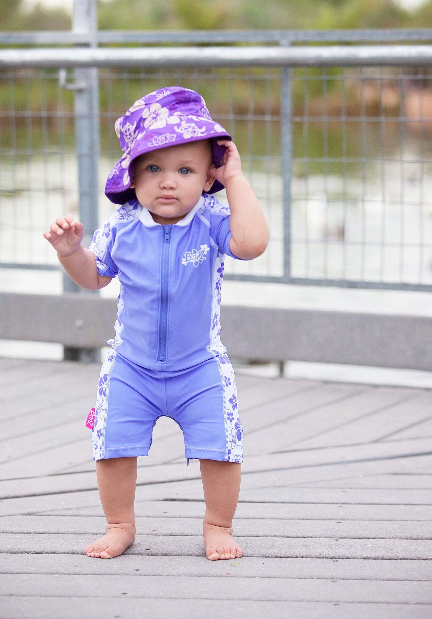 Young child wearing banz one pice zip up swimsuit and reversible sunhat staning on a dock near water