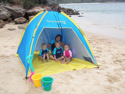 BANZ Global Umbrella Shelta Sun Shelter UV Tent - four children sit inside the tent, buckets and a shovel in the sand outside - they are beachside with rocky cliffs and boats behind