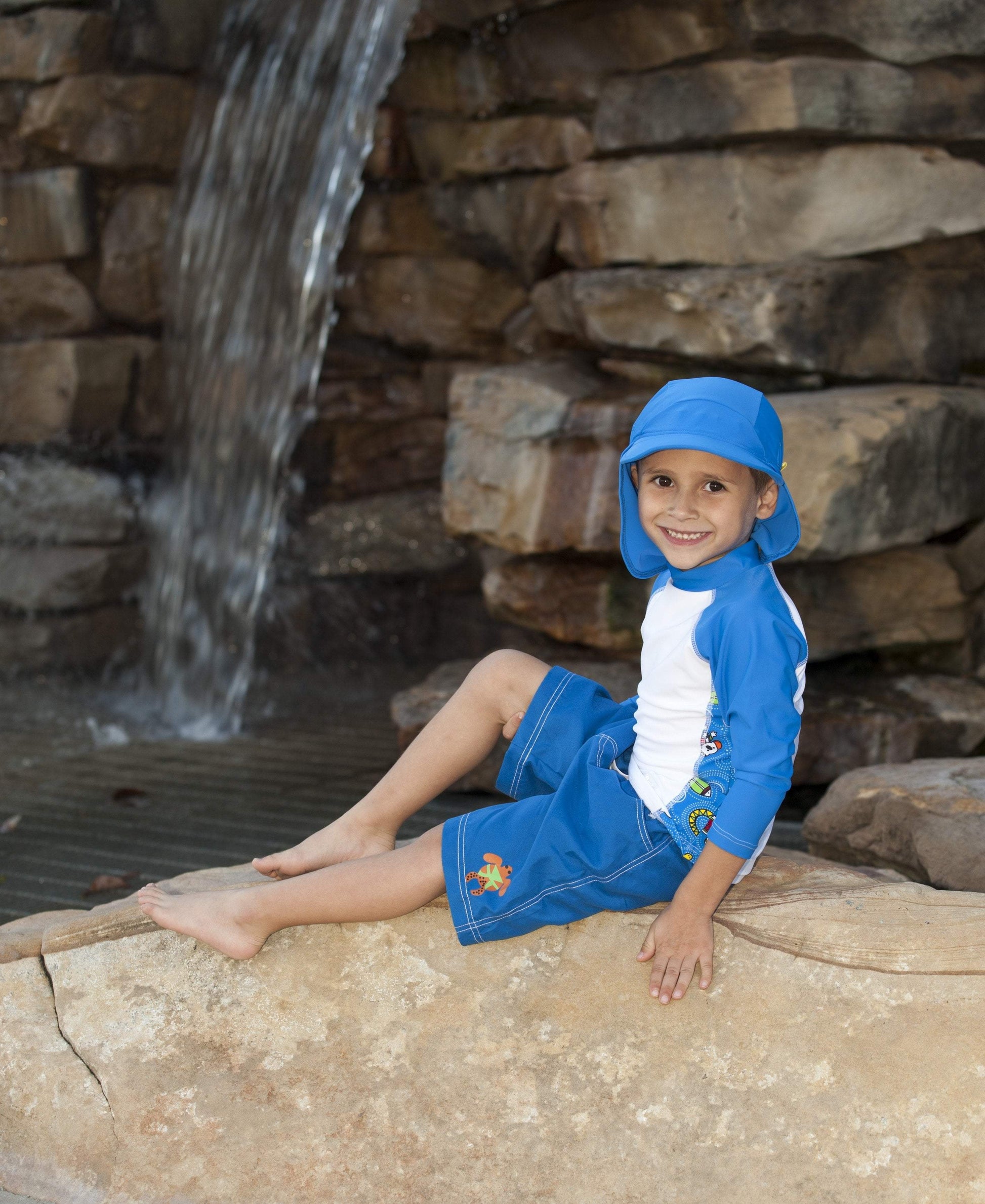 Young child sits near a waterfall and smiles while wearing banz flap hat, long sleeve rash guard and swim trunks