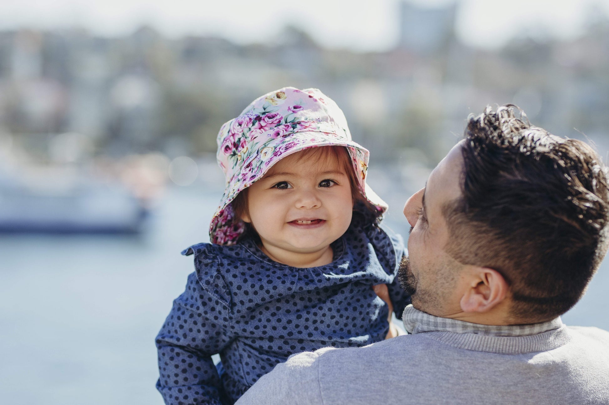 a man holds a young child who smiles as she wears a banz bucket hat near the harbour which is blurred in the background