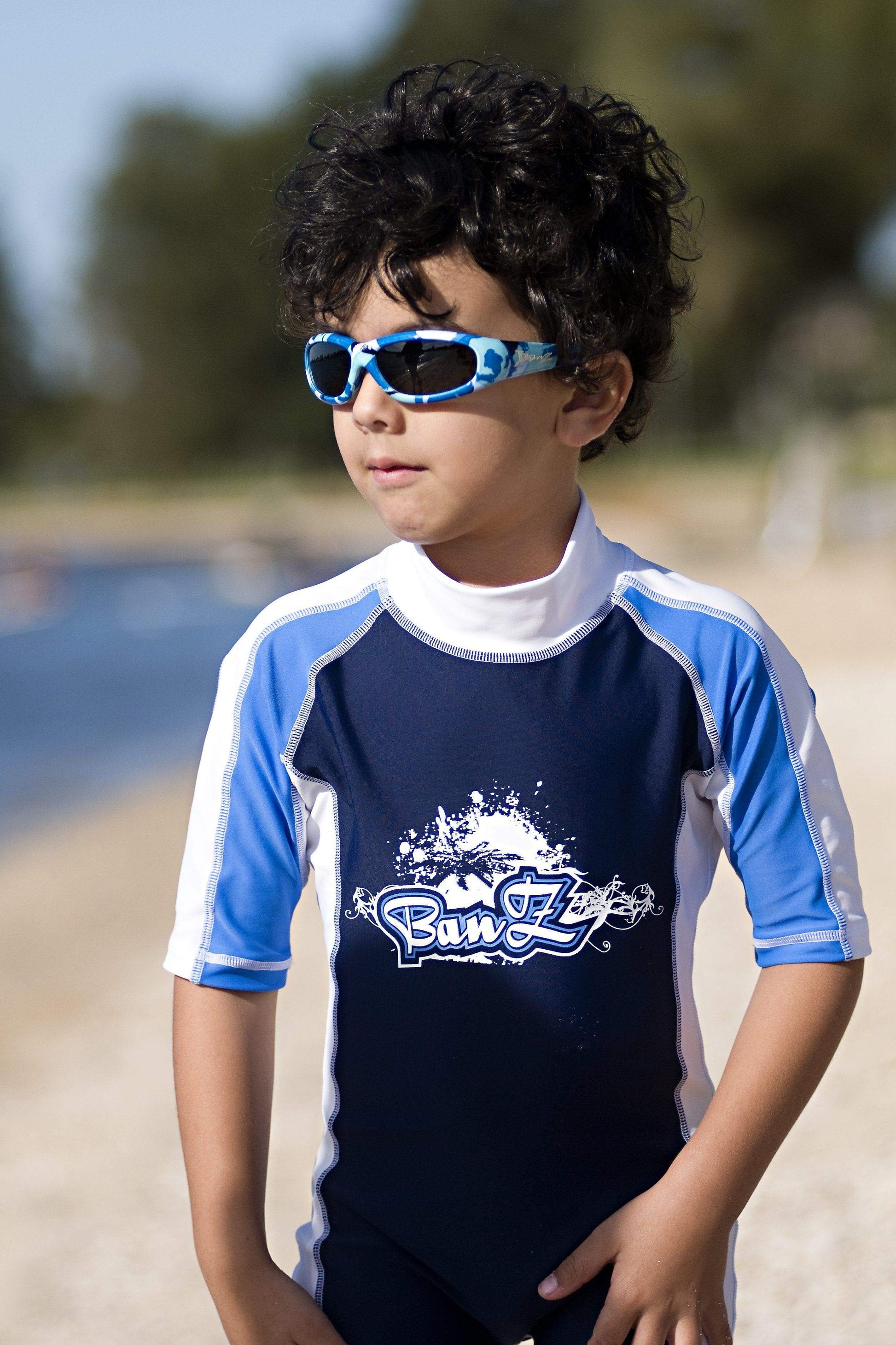 schoolaged child wears banz sunglasses and short sleeved rashtop while at beach