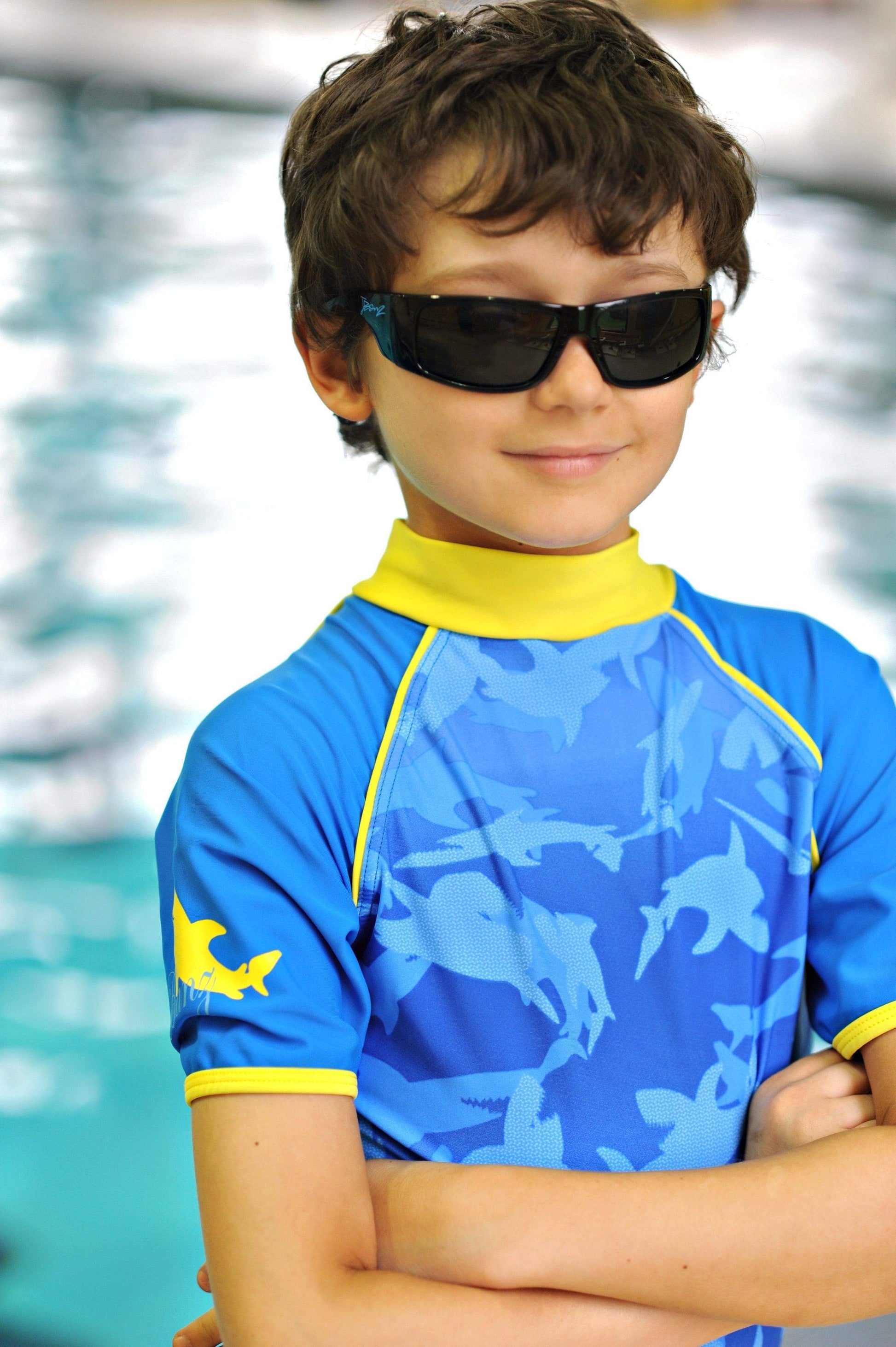 a school age boy crosses his arms and smiles at a pool weaering banz sunglasses and short sleeve rash top