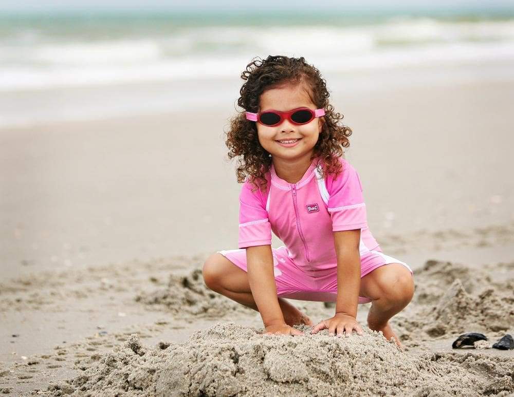 a young child in banz one piece swimsuit abd wrap around sunglasses plays in the sand at the beach